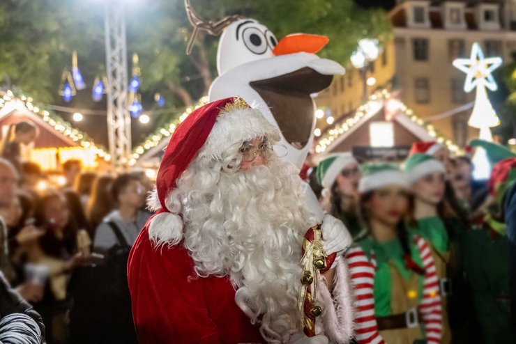 Pai Natal num mercado de Natal iluminado, rodeado de ajudantes vestidos de elfos e um grande boneco de neve decorativo.